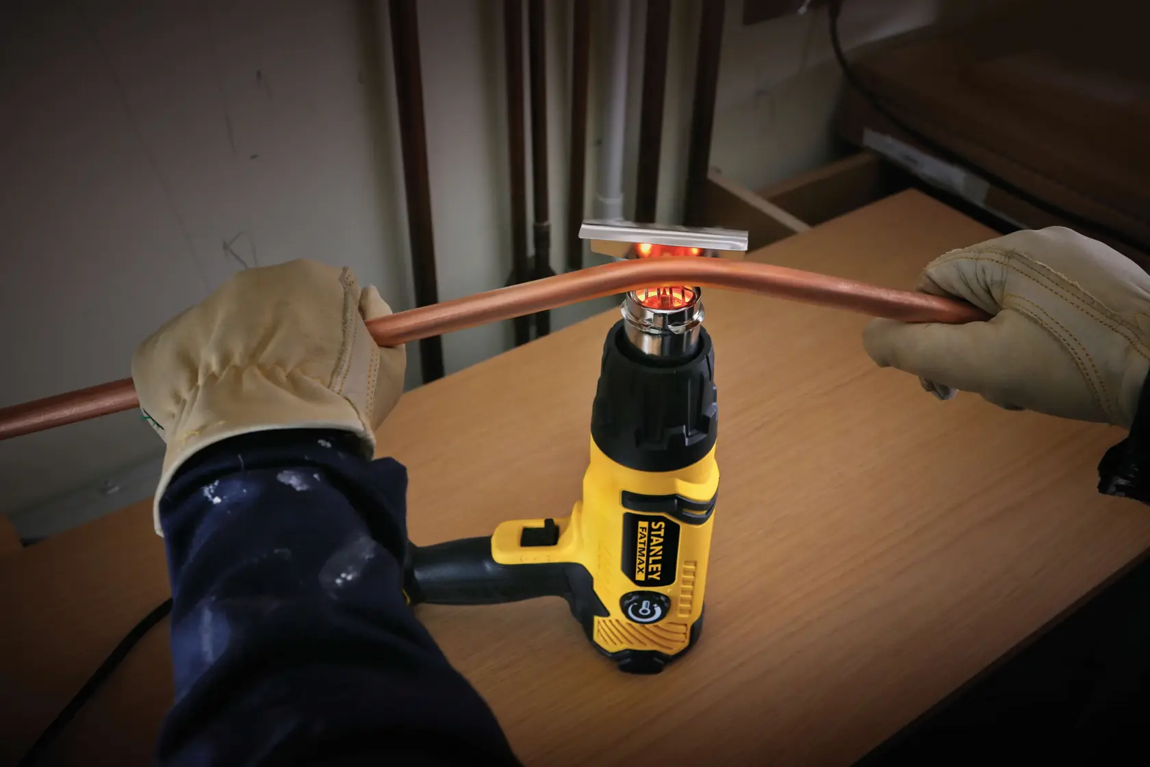 STANLEY FME670K heat gun in use bending a copper pipe, person wearing gloves holds pipe above tool on a workbench.