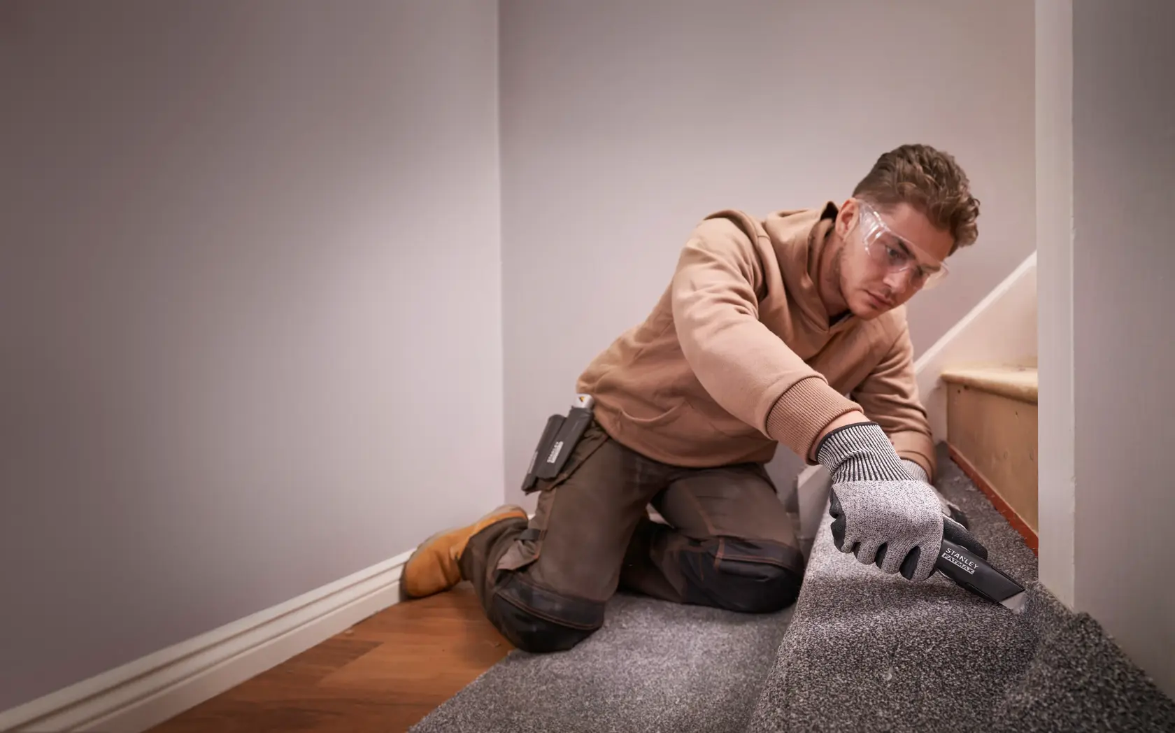 Photo of worker using STANLEY FMHT10500-0 utility knife to cut carpet on stairs, wearing gloves and workwear.