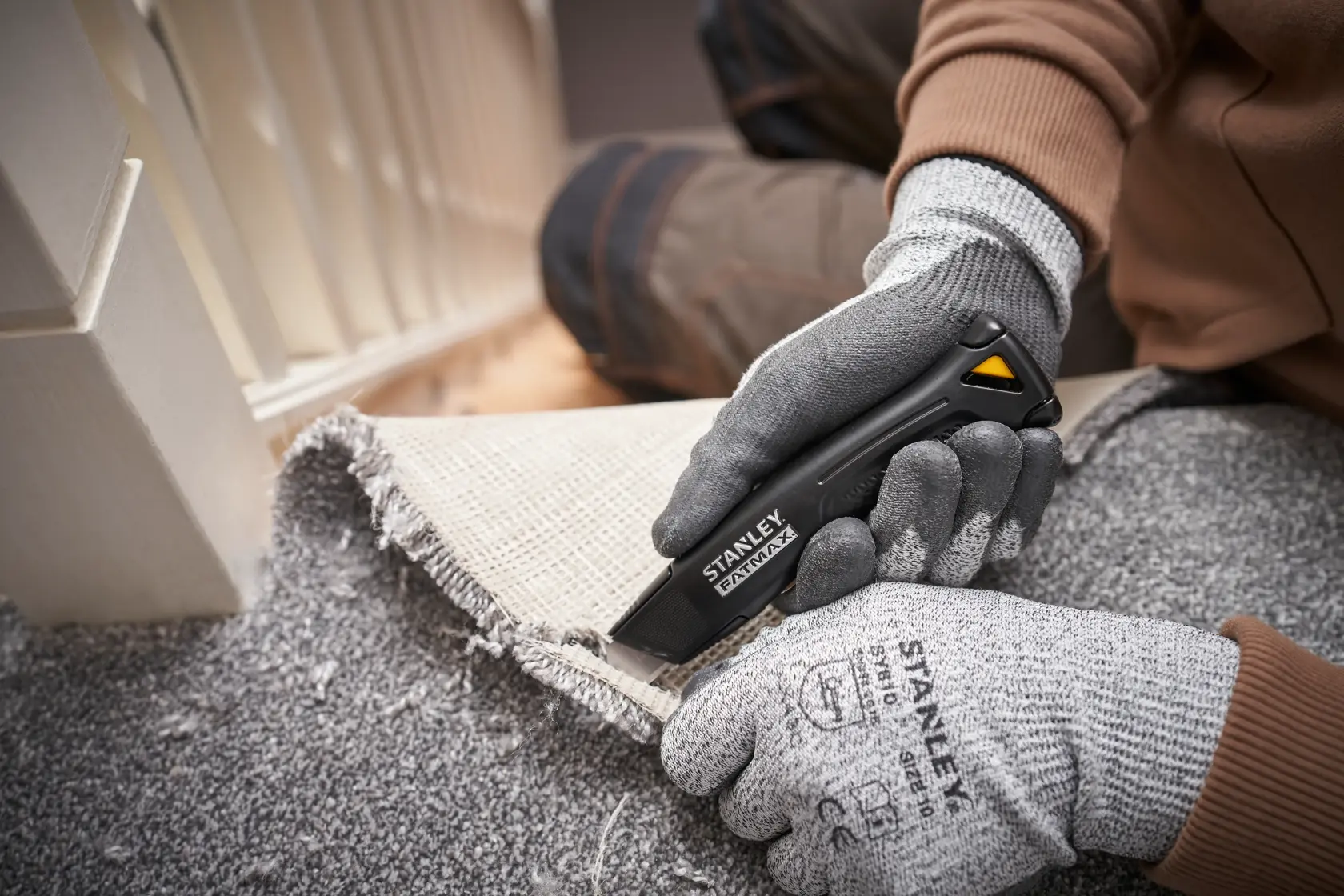 Photo of STANLEY FMHT10500-0 utility knife cutting carpet, gloved hands holding knife and carpet edge.