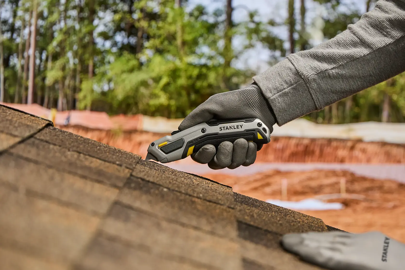 A gloved hand is using a Stanley utility knife to cut roofing shingles on a rooftop, with trees and construction materials in the background.