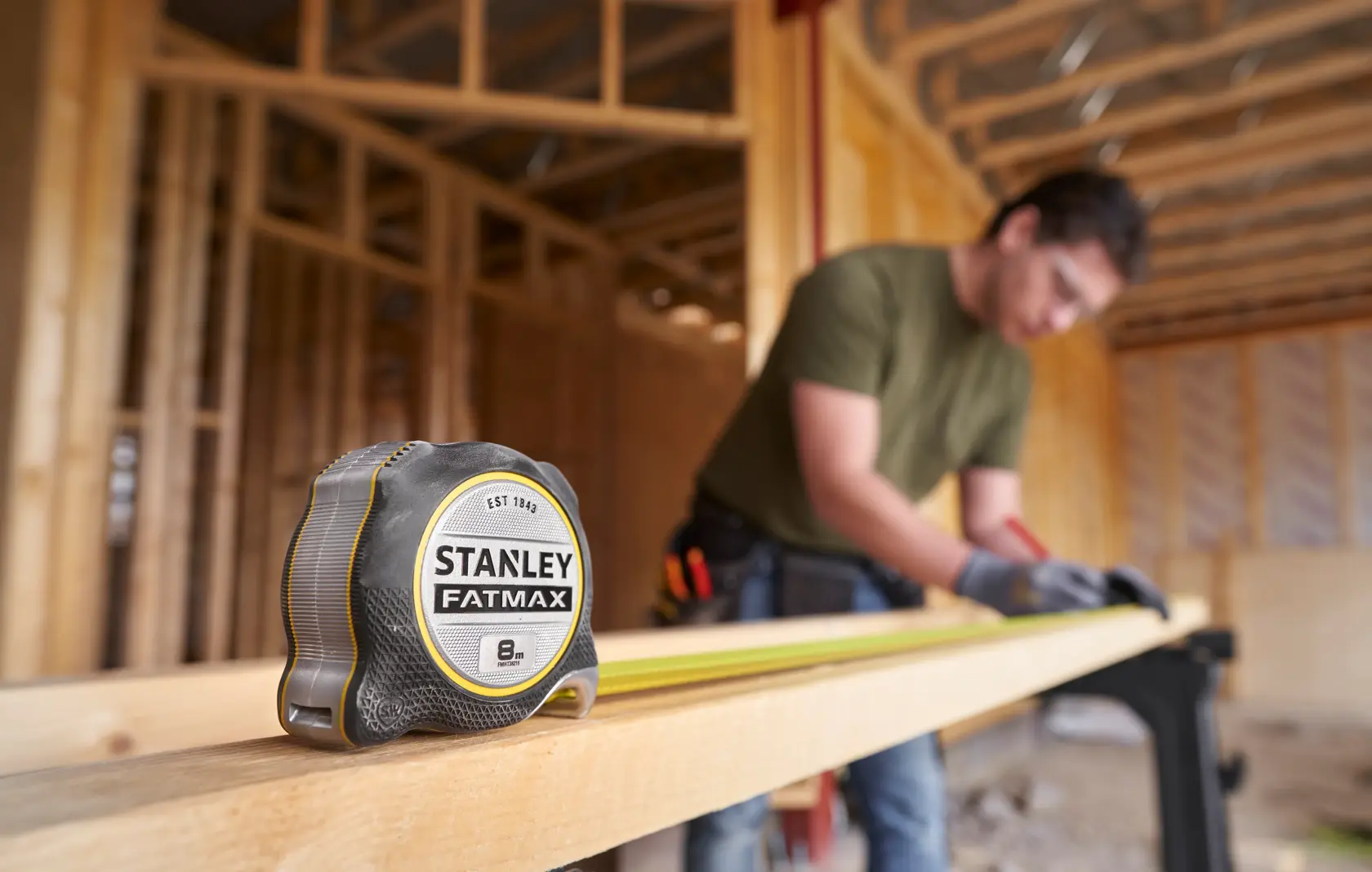 STANLEY FMHT38219-3 tape measure on wooden beam at construction site, blurred worker in background.