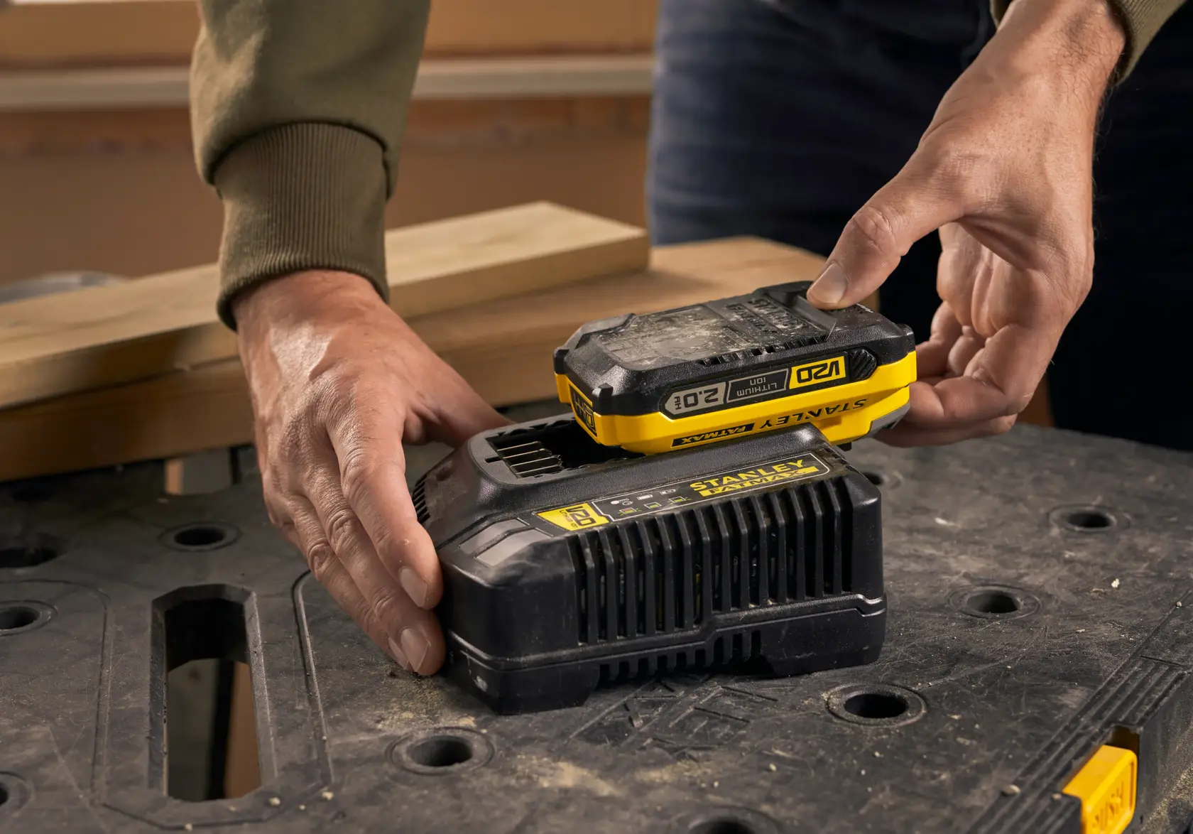 STANLEY SFMCB202 battery being placed on black charger, close-up with workbench in background.