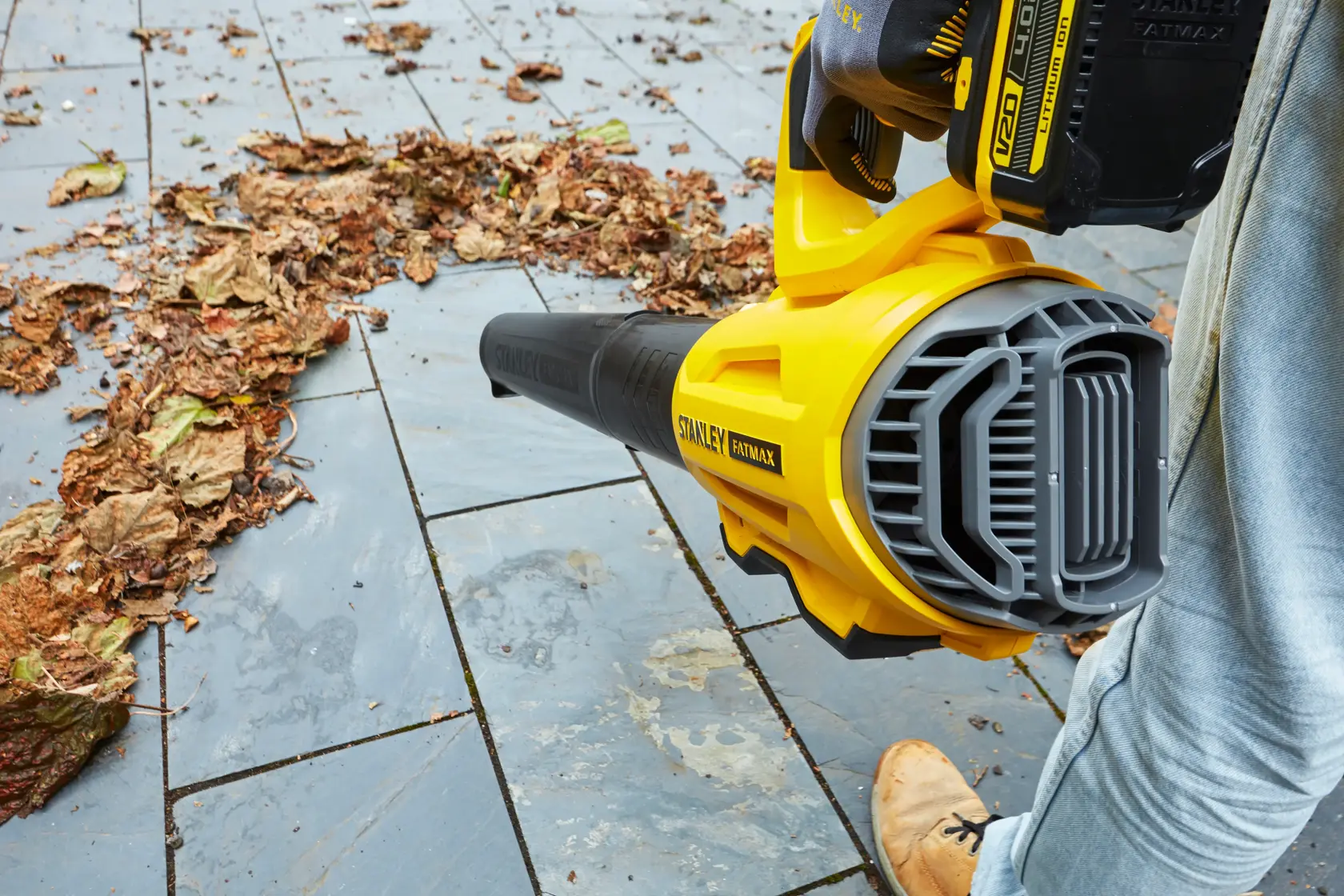 STANLEY SFMCBL7M1 cordless leaf blower close-up, yellow and black, blowing leaves on stone tiles.