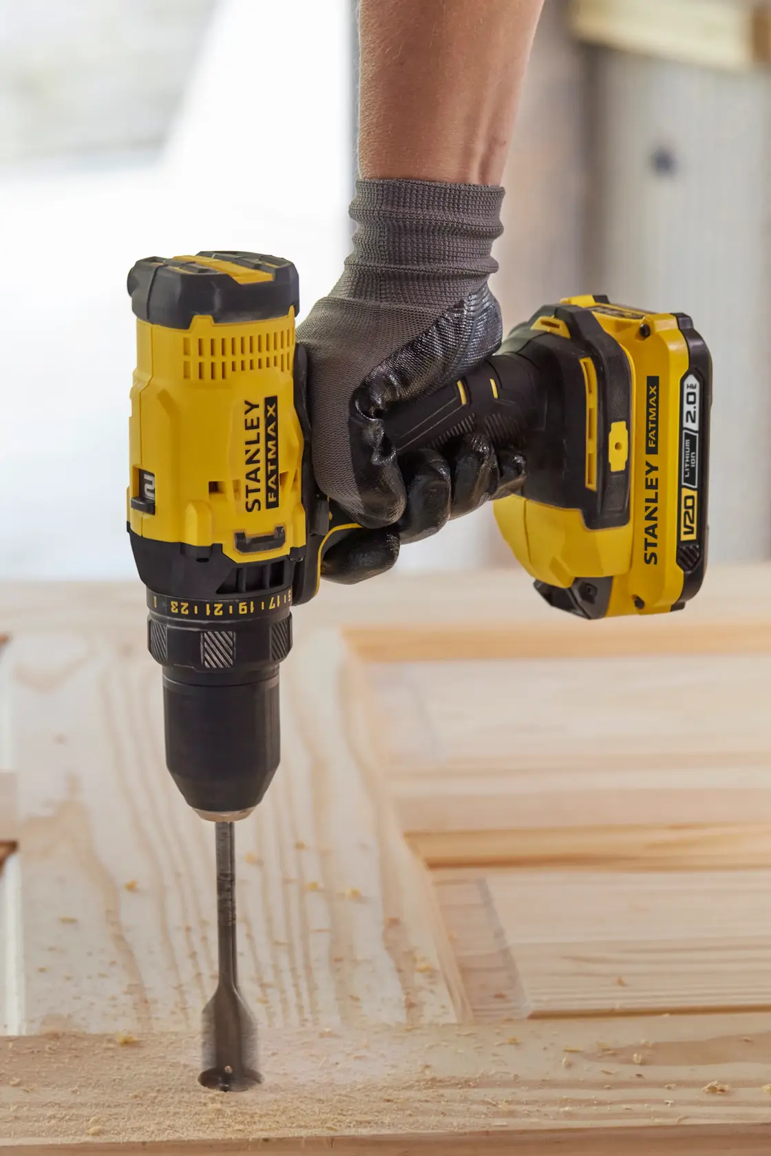 Photo of STANLEY SFMCD700D2S cordless drill driving a spade bit into wood, shown in use with a gloved hand.