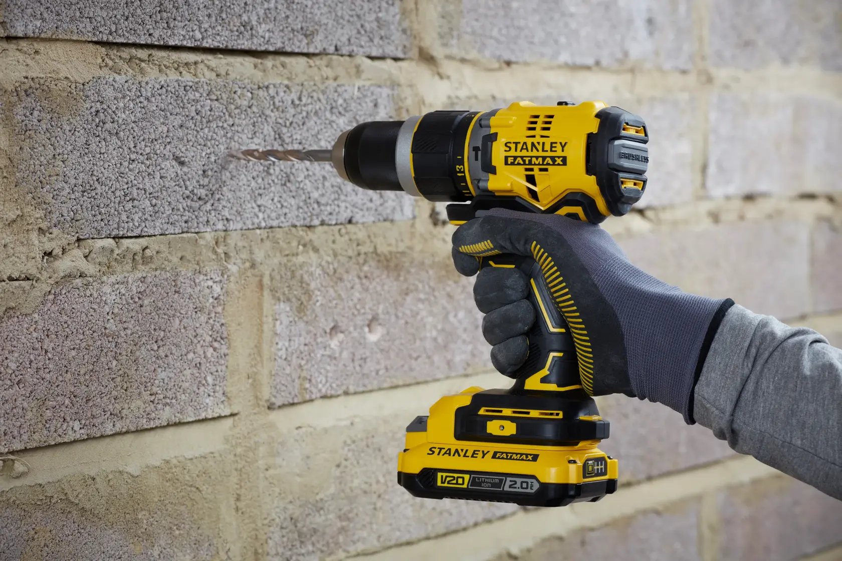 Stanley SFMCD721DR2 cordless drill held against brick wall, yellow and black design, close-up side view.