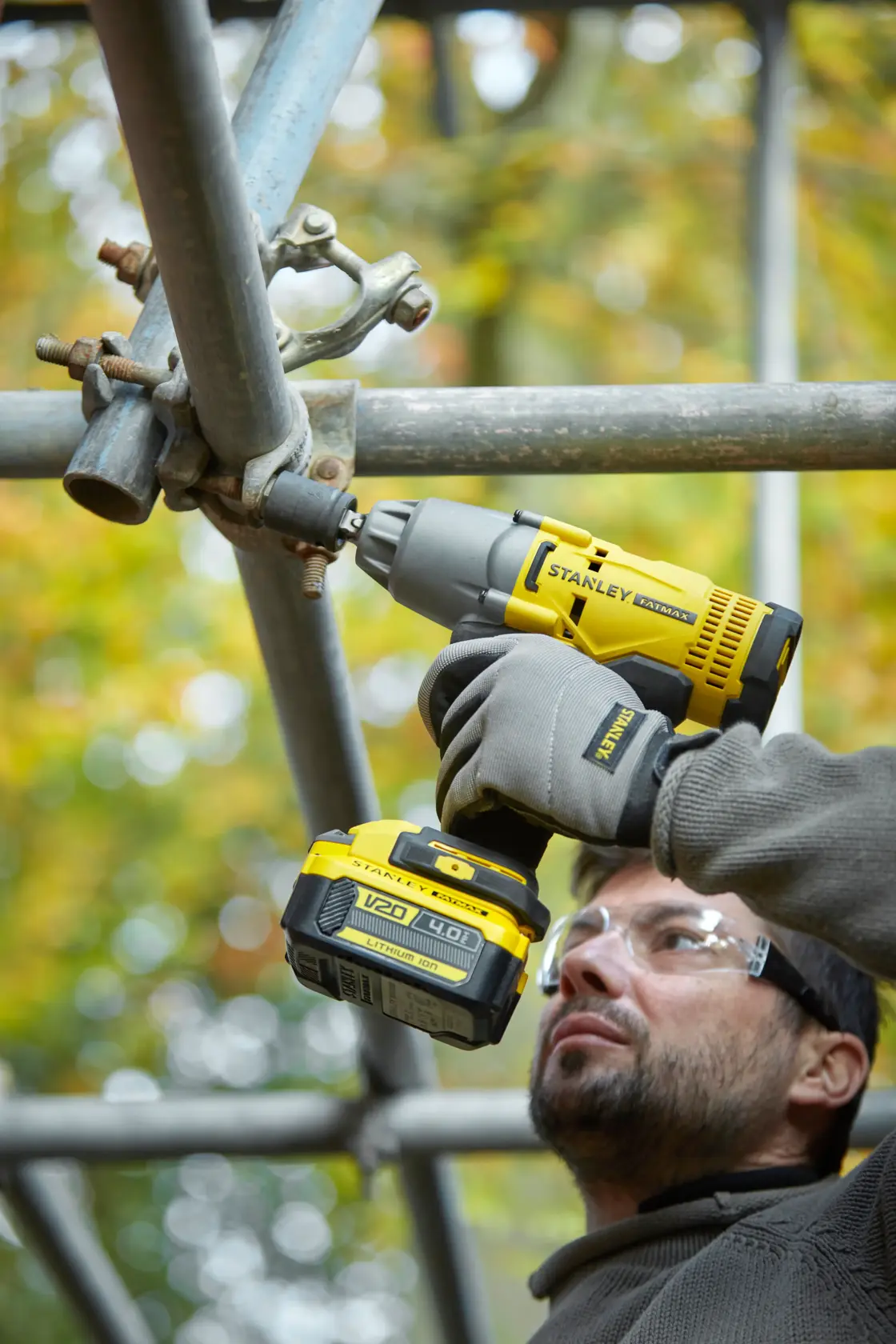 Photo of STANLEY SFMCF900B impact wrench fastening scaffold pipes, held by gloved hands outdoors.