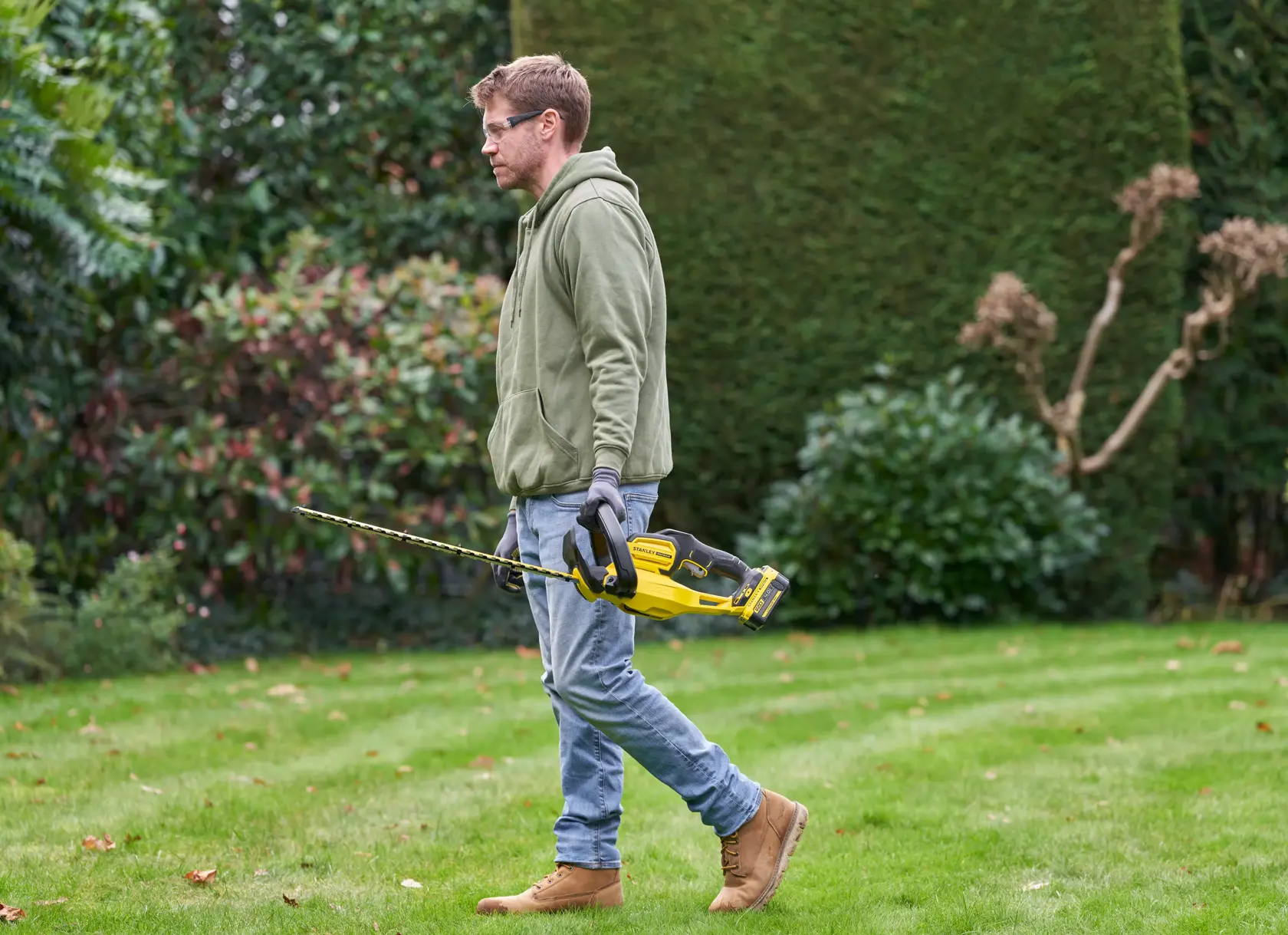 Photo of STANLEY SFMCHT655M1 hedge trimmer held by person in garden, featuring yellow and black design.