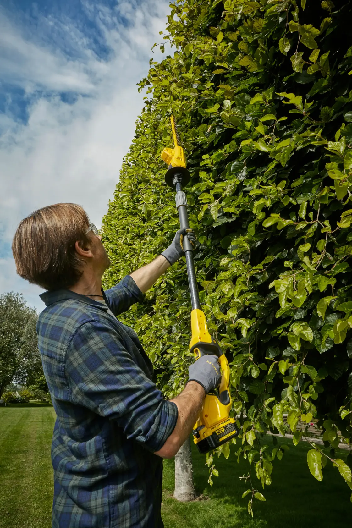 Photo of STANLEY SFMCPH845B pole hedge trimmer in use trimming tall green hedges outdoors.