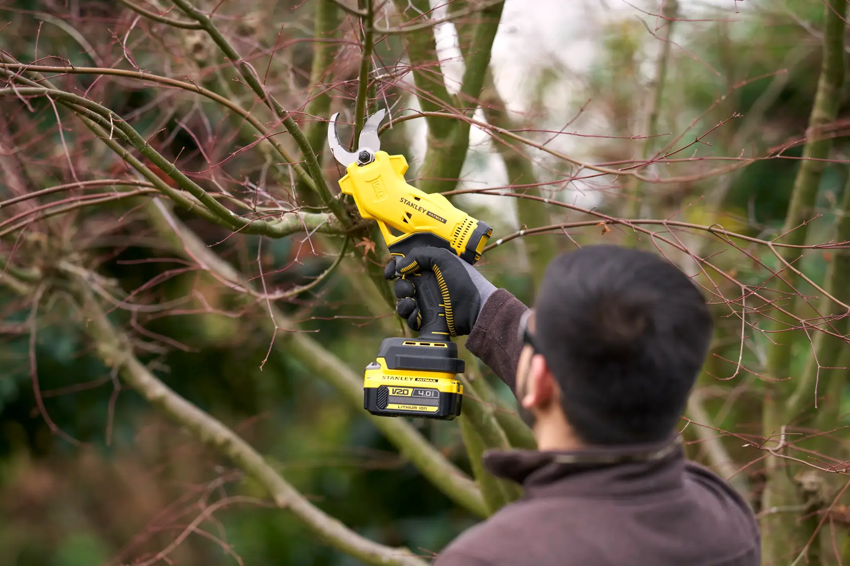 Stanley SFMCPP32B cordless pruning shears in use outdoors, trimming tree branches, product held by gloved hand.