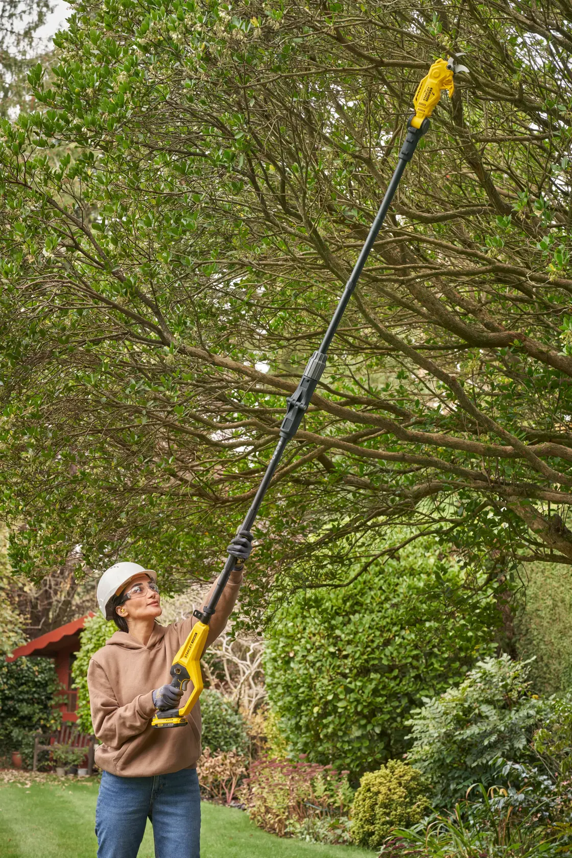 STANLEY SFMCPP632B pole saw in use pruning high tree branches in a garden setting.