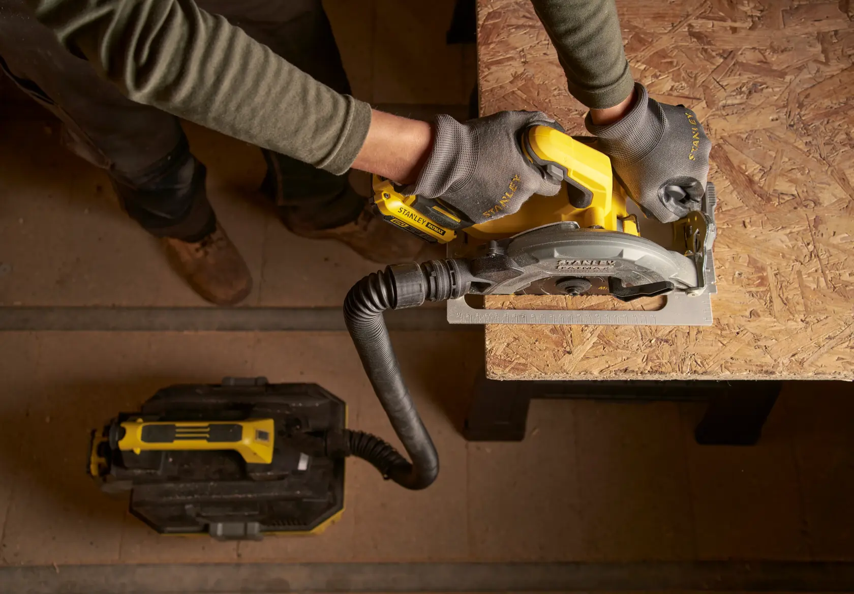Top view of a person using a STANLEY SFMCS550B cordless circular saw to cut wood, attached to a dust extractor.