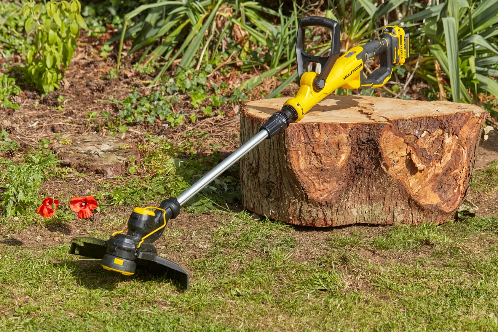 STANLEY SFMCST933B cordless grass trimmer in yellow and black lying on grass near a tree stump outdoors.