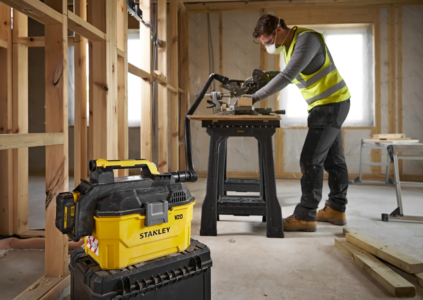 Stanley V20 vacuum in foreground on a toolbox, worker in safety vest using saw in unfinished room.