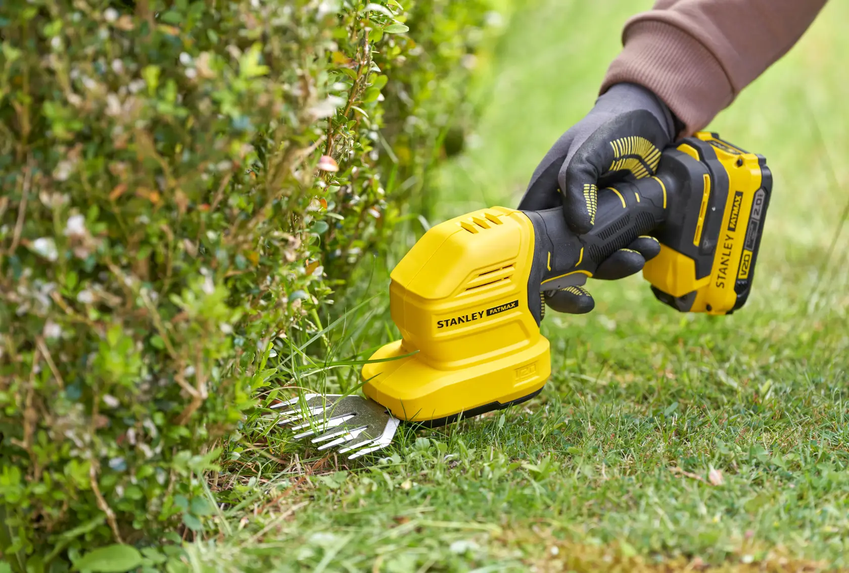 STANLEY STCSS20B hedge grass shear being used to trim grass at the edge of a bush, close-up outdoor view.
