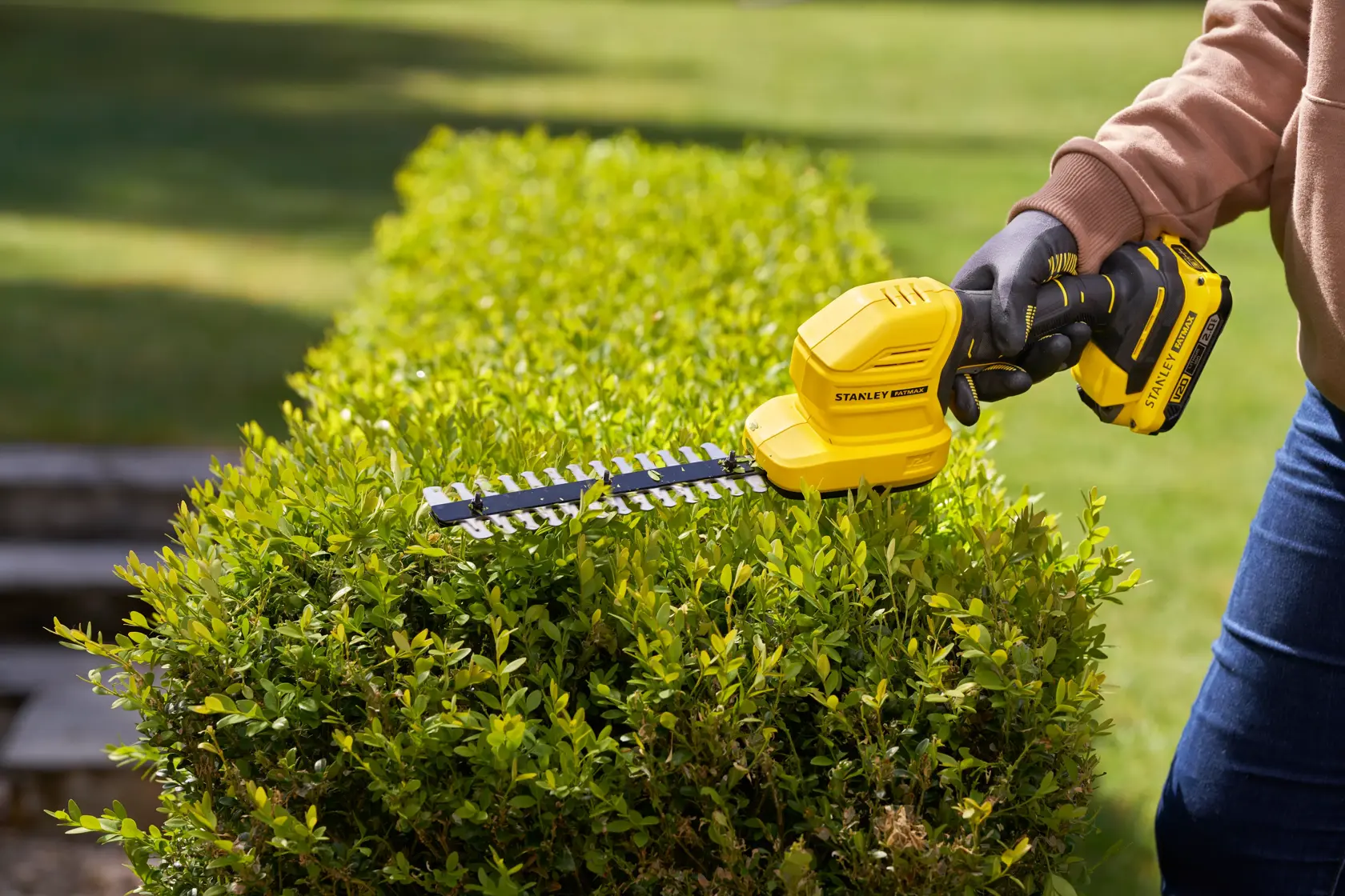 STANLEY STCSS20D1 yellow cordless hedge trimmer in use trimming green bushes outdoors.