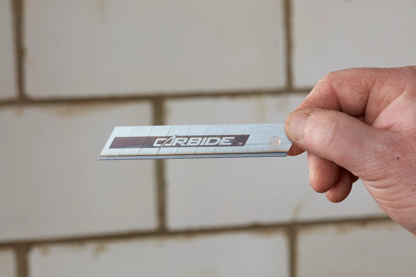 Photo of STANLEY STHT0-11818 carbide utility blade held in hand, sharp edge visible against tile background.