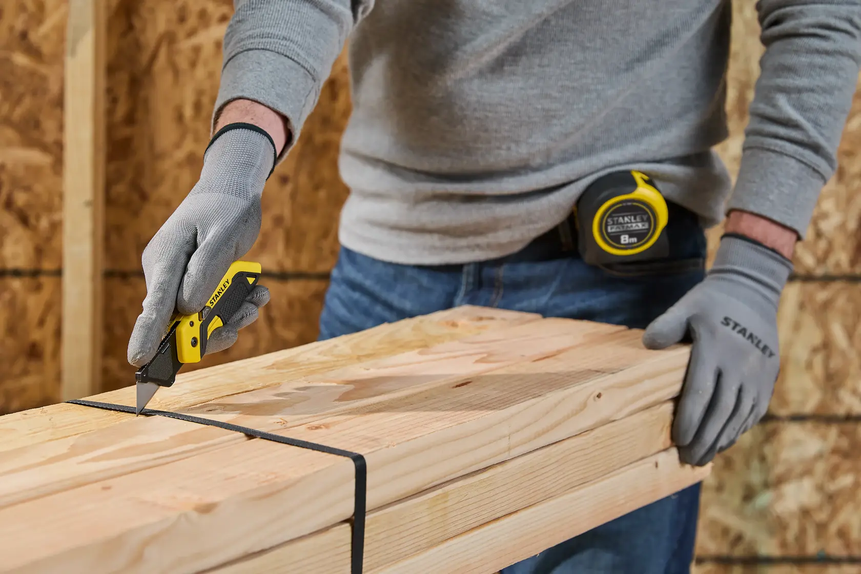 A person wearing grey gloves uses a yellow Stanley utility knife to cut a black strap around a bundle of wooden planks. A Stanley tape measure is clipped to the person's belt.