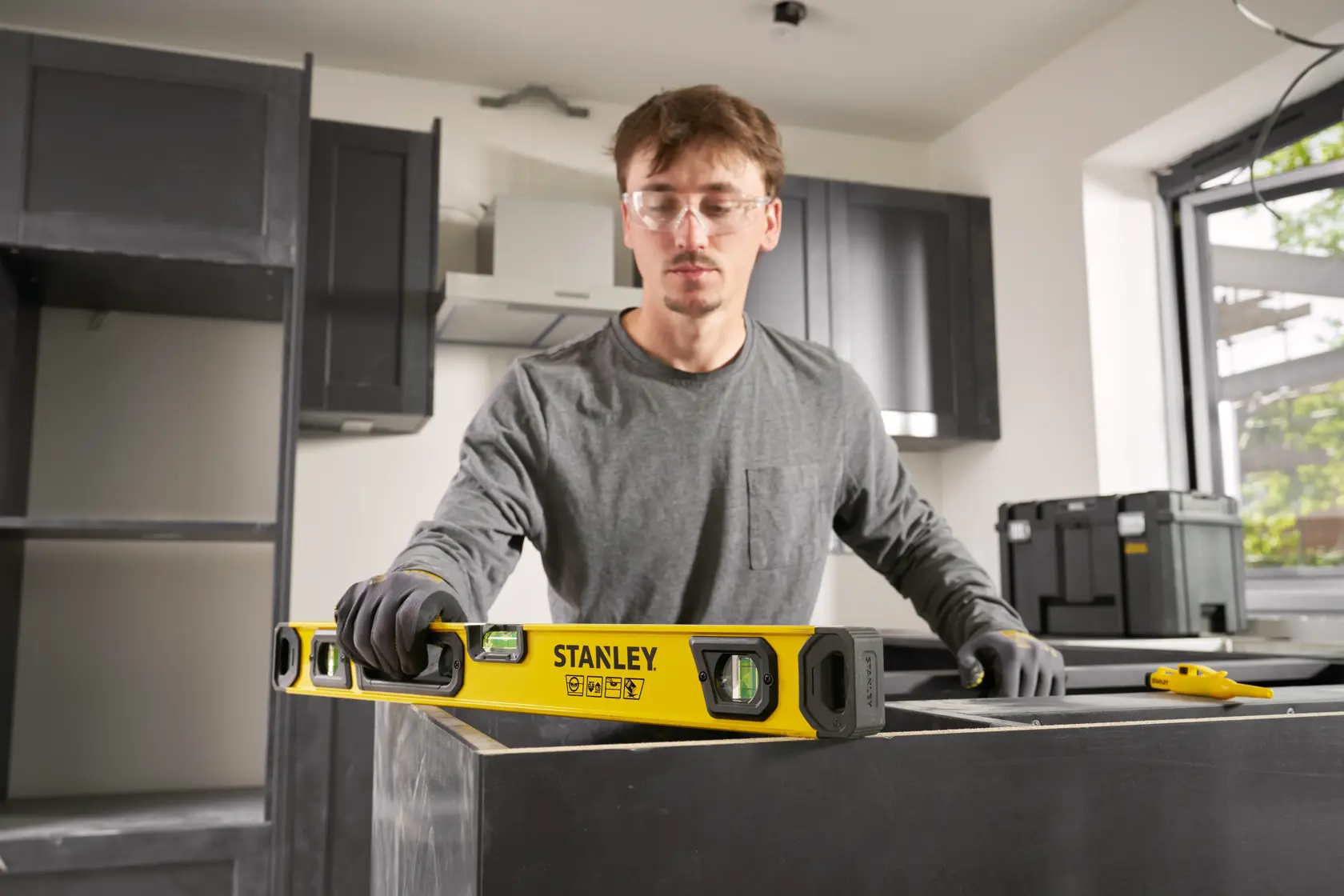 A person wearing gloves uses a yellow STANLEY spirit level to check the horizontal alignment of a kitchen countertop during installation. The scene is set in a modern kitchen under construction.