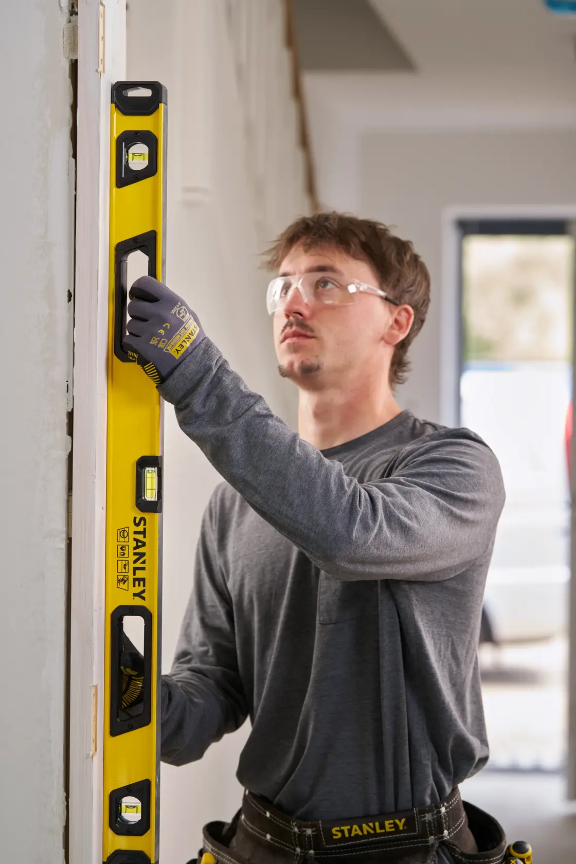 A person wearing work gloves and a tool belt uses a yellow STANLEY spirit level to check the vertical alignment of a door frame inside a building.