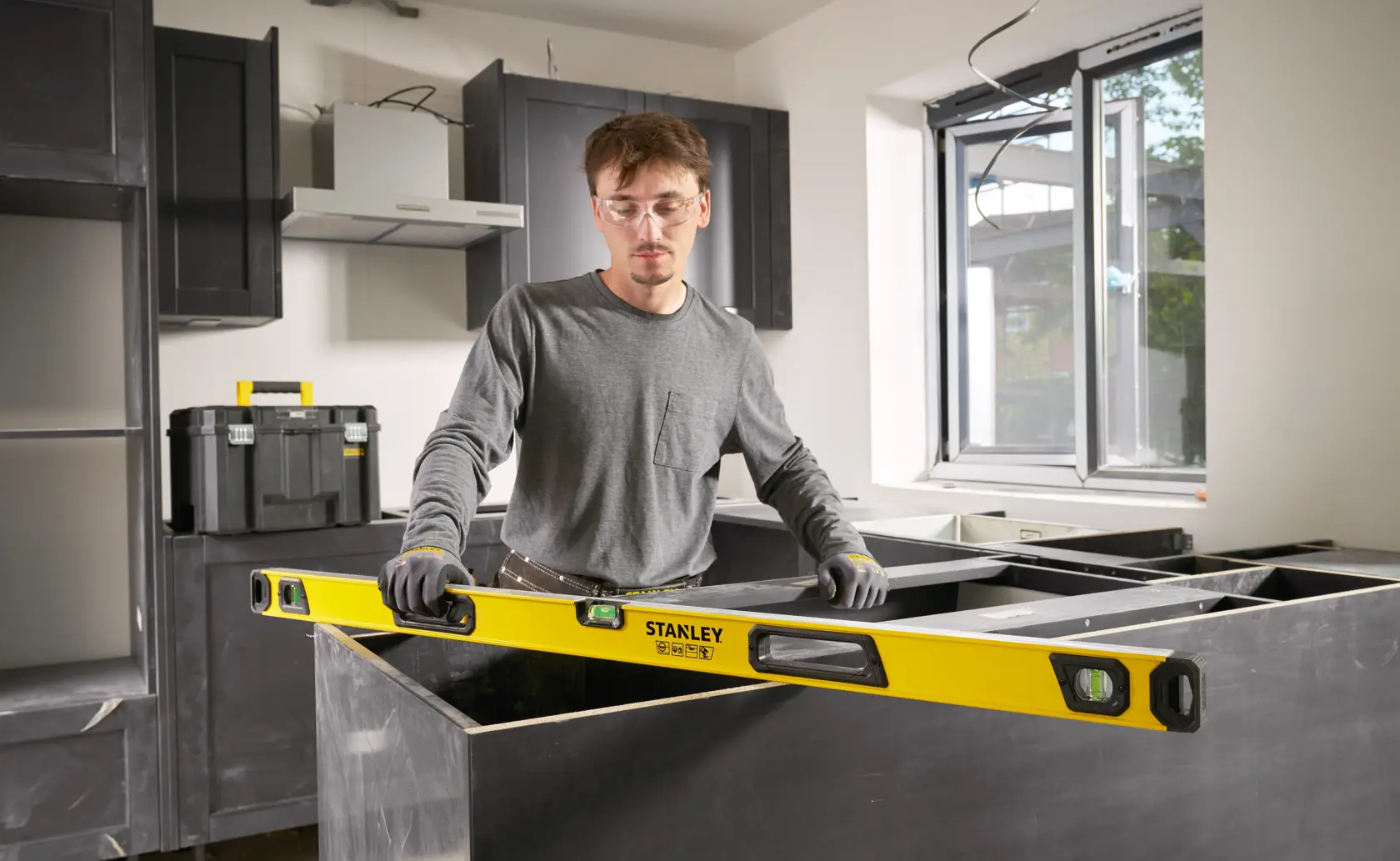 A person with a blurred face is using a STANLEY spirit level (model STHT41048-0) to check alignment during construction or installation in a modern kitchen. The spirit level is yellow with black accents and has visible bubble vials. A toolbox is seen in the background.
