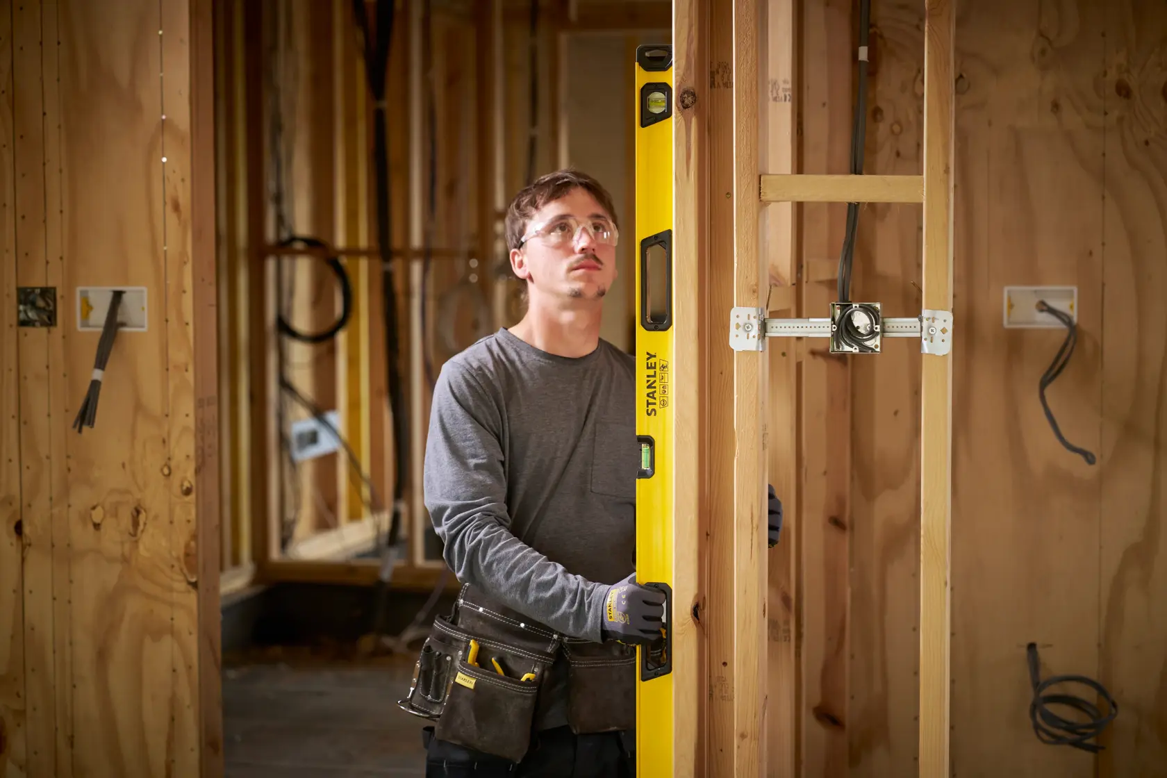 A person with their face blurred is holding a yellow STANLEY spirit level inside a wooden frame of a house under construction. The person is wearing a tool belt and gloves, and electrical wiring is visible in the wall.